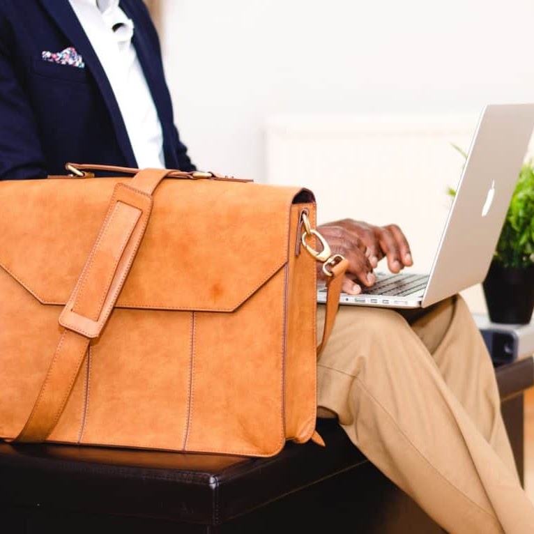 person sitting and typing on a laptop with a leather bag next to them